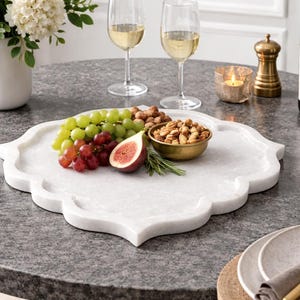 Puede incluir: Una bandeja de mármol blanco con borde decorativo contiene uvas verdes y rojas, higos y nueces. Un pequeño cuenco dorado contiene nueces. Copas de vino, una vela y una botella de vino están sobre una mesa gris.