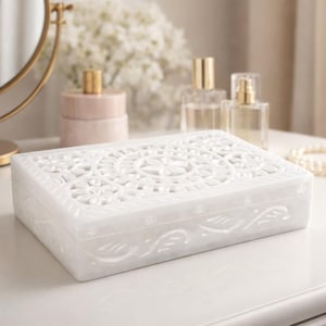 Puede incluir: Una caja de joyería de mármol blanco con intrincados patrones florales tallados en la tapa y los lados. La caja rectangular está sobre una superficie blanca, junto a un labial, frascos de perfume y un espejo.