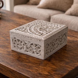 Op de afbeelding: Een rechthoekige, lichtgrijze decoratieve doos met ingewikkelde gebeeldhouwde bloemen- en geometrische patronen. De doos is gemaakt van steen en staat op een donkerbruine houten tafel. Het deksel is ook gesneden.