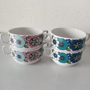 Op de afbeelding: Vier witte keramische soepkommen met handvatten, in paren gestapeld. Elke kom heeft een bloemenpatroon in tinten blauw, roze en groen. De kommen staan tegen een effen witte achtergrond.