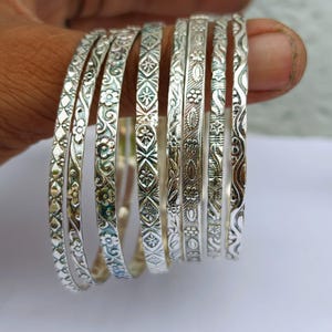 Puede incluir: Un conjunto de siete brazaletes de metal en tono plateado, cada uno con un patrón en relieve distinto. Los diseños incluyen motivos florales, formas geométricas de diamante, líneas onduladas y detalles texturizados en cada pulsera.