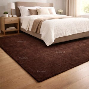 May include: A dark brown rectangular rug in a bedroom setting. The rug is placed on a wooden floor in front of a bed with white bedding and a tan headboard. The rug has a textured appearance and is labeled with the text "FAMRUG".