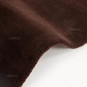 May include: Close-up of a dark brown rug with a soft, velvety texture. The rug has a slightly curved edge and a visible seam. The brand name "FAMRUG" is visible in the corners. The rug appears to be made of a plush material.