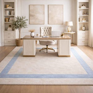 May include: An elegant home office setup featuring a large beige rug with a light blue border. A wooden desk with cream-colored panels and a tufted chair are centered. Two matching bookshelves flank the desk, creating a sophisticated workspace.