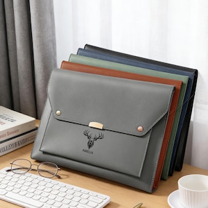 May include: A selection of leather document holders in shades of grey, brown, and black. The front grey holder displays a deer graphic and the name "AMELIA". Each holder has a flap closure with gold-coloured hardware.