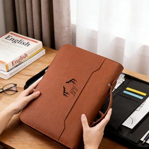 May include: A brown portfolio with a textured surface, featuring a black emblem. The portfolio has a handle and is open to reveal a black interior with card slots and documents. English books and eyeglasses are visible in the background.