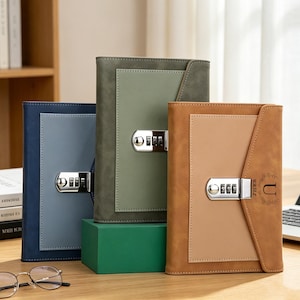 Könnte beinhalten: Drei ledergebundene Notizbücher in Blau, Grün und Braun, jeweils mit Zahlenschloss. Die Notizbücher haben ein zweifarbiges Design mit einem helleren rechteckigen Feld auf der Vorderseite. Das braune Notizbuch hat den Buchstaben "U" und das Wort "U.R.J.E" aufgedruckt.