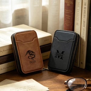 May include: Two leather card wallets with zip closures. One is brown with a dragon graphic and the name "BECK". The other is black with the name "McGinn" and the initial "M". Both have a small pocket on the front.