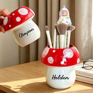 May include: A red and white mushroom-shaped ceramic makeup brush holder with the name "Holden" in black script. The holder contains several makeup brushes. A matching mushroom-shaped cup with the name "Grayson" is held nearby.