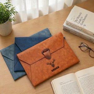 May include: Three leather document holders in shades of blue and brown are displayed on a wooden surface. The brown holder features a buckle closure and the word "XAVIER" with a deer graphic. Books, glasses, and a potted plant are also visible.