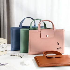 May include: Three tote bags in blue, green, and pink, with a brown bag in the foreground. The bags have handles and a flap closure with a button. The pink bag has a pen illustration and the name "William".