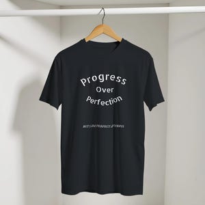Könnte beinhalten: Ein schwarzes T-Shirt mit kurzen Ärmeln und weißem Text, der in geschwungener Schrift Progress Over Perfection lautet, sowie dem kleineren Schriftzug BUT I AM PERFECT ANYWAYS darunter. Das Kleidungsstück hängt an einem Holzkleiderbügel.
