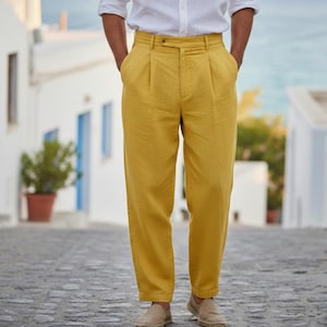 Peut inclure: Pantalon plissé jaune moutarde avec fermeture boutonnée et poches. Le pantalon est associé à des espadrilles beiges. La personne porte une chemise blanche boutonnée. En arrière-plan, des bâtiments blancs et une rue pavée.