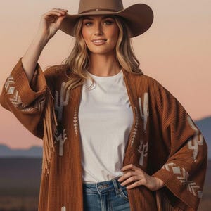 Puede incluir: Una mujer con un sombrero de vaquero marrón y una rebeca color óxido con bordados de cactus blancos. La rebeca tiene flecos en la parte inferior y en las mangas. Lleva una camiseta blanca y vaqueros azules.