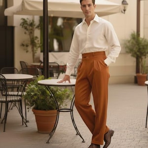 May include: A man in a white shirt and rust-coloured pleated trousers. He is standing near a small round table with a plant in a terracotta pot. He is wearing brown leather shoes. The trousers are a burnt orange colour.
