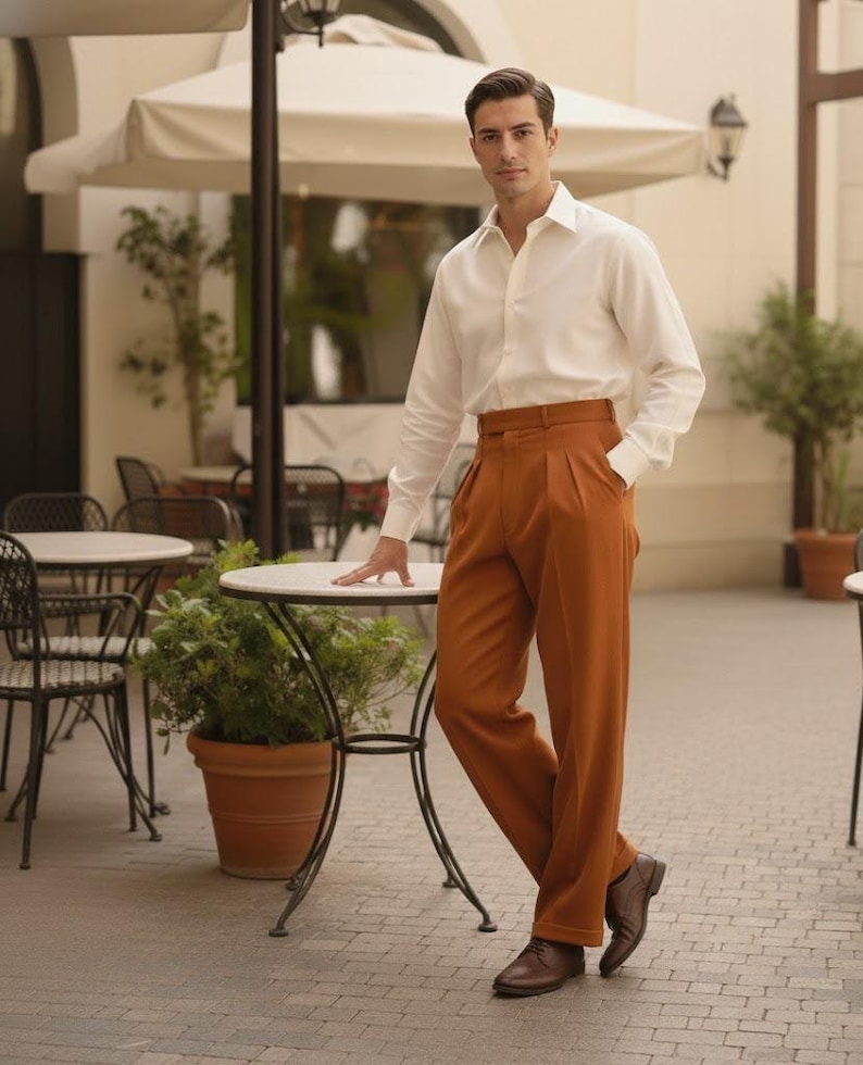 May include: A man in a white shirt and rust-coloured pleated trousers. He is standing near a small round table with a plant in a terracotta pot. He is wearing brown leather shoes. The trousers are a burnt orange colour.