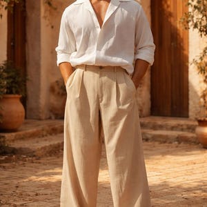 Pode incluir: Uma pessoa vestindo uma camisa de linho branca com gola aberta e calças bege de perna larga. As calças têm pregas na cintura. A pessoa está usando sapatos brancos. O fundo é um edifício de pedra com portas de madeira.