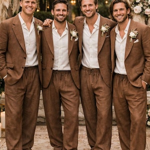 May include: Four men in matching brown suits, white shirts, and brown shoes stand together. Each suit includes a jacket and drawstring pants. They wear small white flower boutonnieres. The setting appears to be an outdoor wedding.