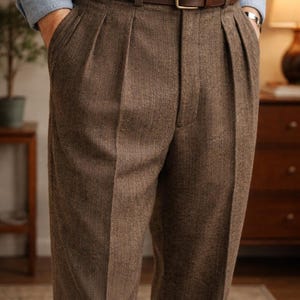 Puede incluir: Pantalones marrones con pliegues con cinturón de cuero marrón a juego y hebilla dorada. Los pantalones tienen un patrón de espiga y se usan con una camisa abotonada azul claro. El atuendo es clásico y elegante.