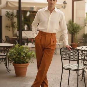 May include: A person in a white shirt and rust-coloured, high-waisted trousers with pleats. They are wearing brown shoes. The scene is set in an outdoor cafe. The trousers are a shade of brown.