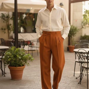 May include: A man in a cream-coloured button-down shirt and rust-coloured pleated trousers. The trousers have a high waist and a belt. He is wearing brown leather shoes. The background includes outdoor seating and potted plants.