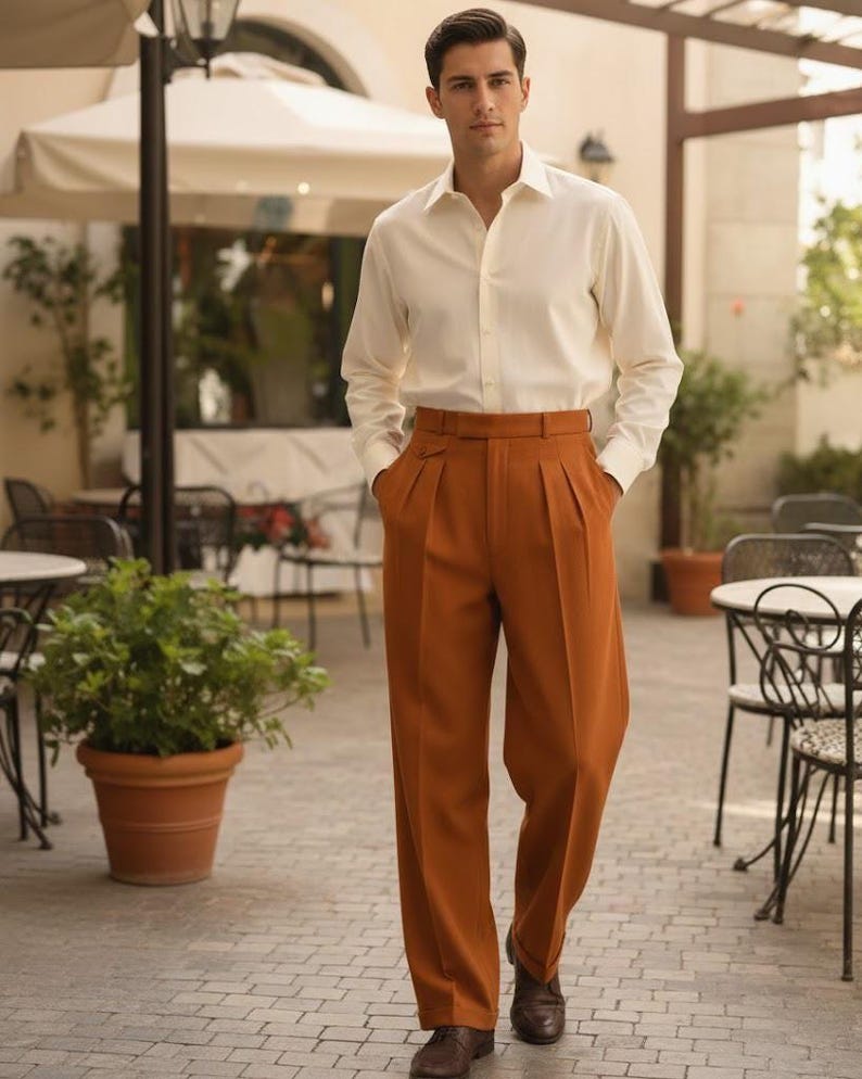 May include: A man in a cream-coloured button-down shirt and rust-coloured pleated trousers. The trousers have a high waist and a belt. He is wearing brown leather shoes. The background includes outdoor seating and potted plants.