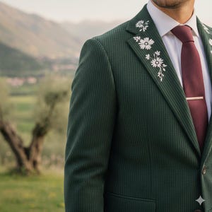 Può includere: Blazer in velluto a coste verde scuro con ricami floreali bianchi sul bavero. Il blazer è abbinato a una cravatta bordeaux e a un fermacravatta dorato. Lo sfondo presenta un paesaggio sfocato con montagne e alberi.