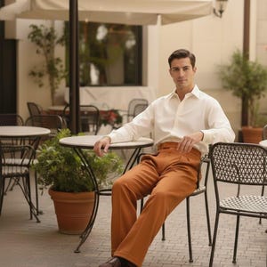 May include: A man in a white shirt and rust-coloured trousers sits at a cafe table. He wears brown leather shoes. The outdoor setting includes a white umbrella and other tables. The trousers are a warm, earthy tone.