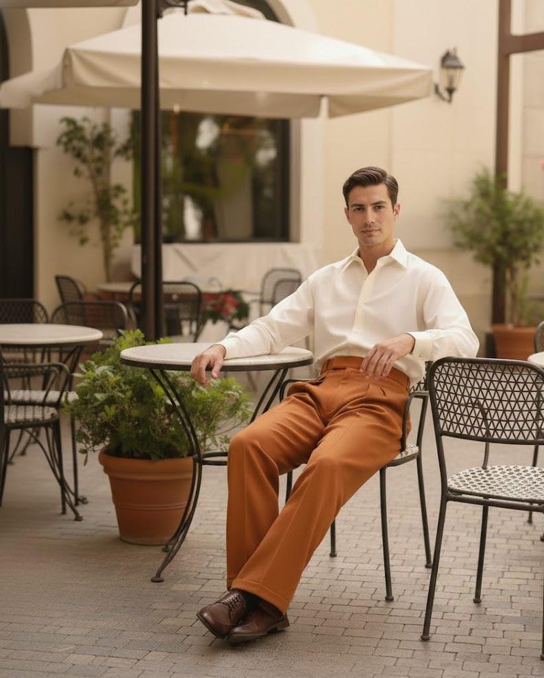 May include: A man in a white shirt and rust-coloured trousers sits at a cafe table. He wears brown leather shoes. The outdoor setting includes a white umbrella and other tables. The trousers are a warm, earthy tone.