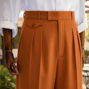 May include: Close-up of rust-coloured pleated trousers with a high waist and a buttoned pocket. The trousers are paired with a white collared shirt with rolled-up sleeves. The fabric appears to be a smooth, woven material.