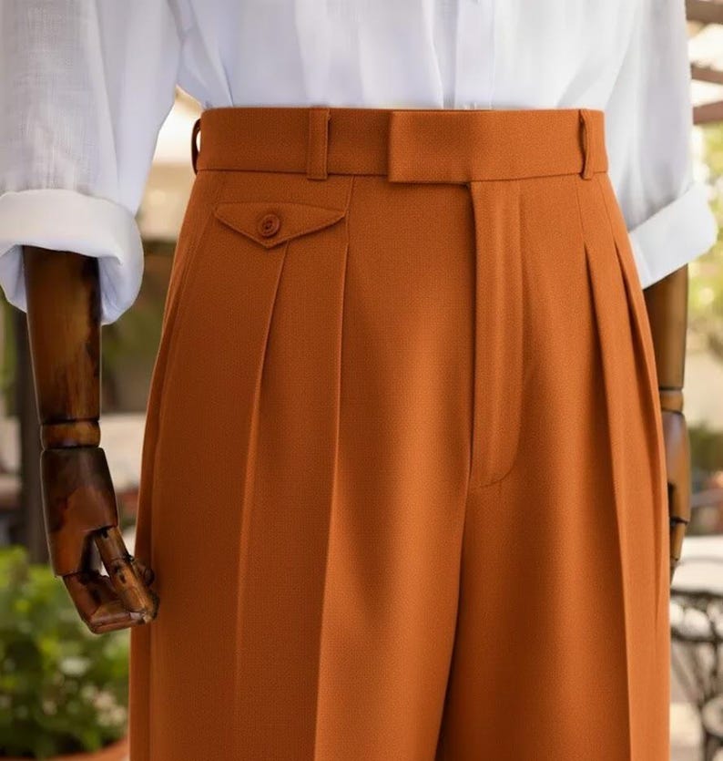 May include: Close-up of rust-coloured pleated trousers with a high waist and a buttoned pocket. The trousers are paired with a white collared shirt with rolled-up sleeves. The fabric appears to be a smooth, woven material.