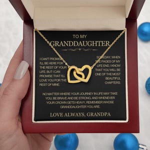 Op de afbeelding: Een goudkleurige ketting met twee in elkaar grijpende hartvormige hangers op een zwarte kaart in een donkerhouten doosje. De kaart bevat gouden tekst gericht aan een kleindochter, waaronder: TO MY GRANDDAUGHTER, LOVE ALWAYS, GRANDPA.