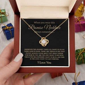 Può includere: Una collana color oro con un ciondolo a nodo e un cristallo rotondo trasparente, presentata in una scatola con un cartoncino nero. Il testo recita: When you wear this Promise Necklace always remember... seguito da un lungo messaggio e I Love You.