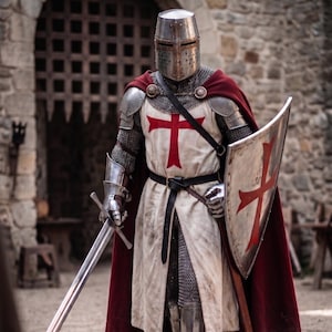 May include: A knight in full armor, including a helmet, chainmail, and metal greaves, stands holding a sword and shield. The knight wears a white tunic with a red cross, a red cape, and a black belt. The shield also features a red cross.
