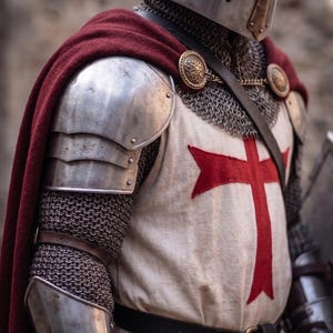 May include: A medieval knight in full armor, including a helmet, chainmail, and plate armor. A red cape is draped over the shoulders. The knight's tunic features a large red cross. The image is a close-up, focusing on the upper body.