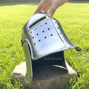Può includere: Un elmo medievale argentato lucido con la parte superiore arrotondata, una fessura per gli occhi e numerosi fori di ventilazione. L'elmo è appoggiato su una pietra in un'area erbosa. Il testo "MedievalForgeMD" è visibile.