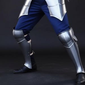 以下が含まれることがあります： 磨かれた金属製の鎧を身につけた人の脚のクローズアップ。 鎧にはすね当てと膝の保護具が含まれており、青いズボンと黒い靴と組み合わされています。 金属は反射面を持っており、歴史的な再現やコスプレを示唆しています。