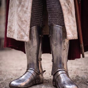 May include: Close-up of medieval steel leg armor, including greaves and sabatons. The armor is highly polished, reflecting the light. The legs are covered in chainmail. A white and red cloak is visible in the background.