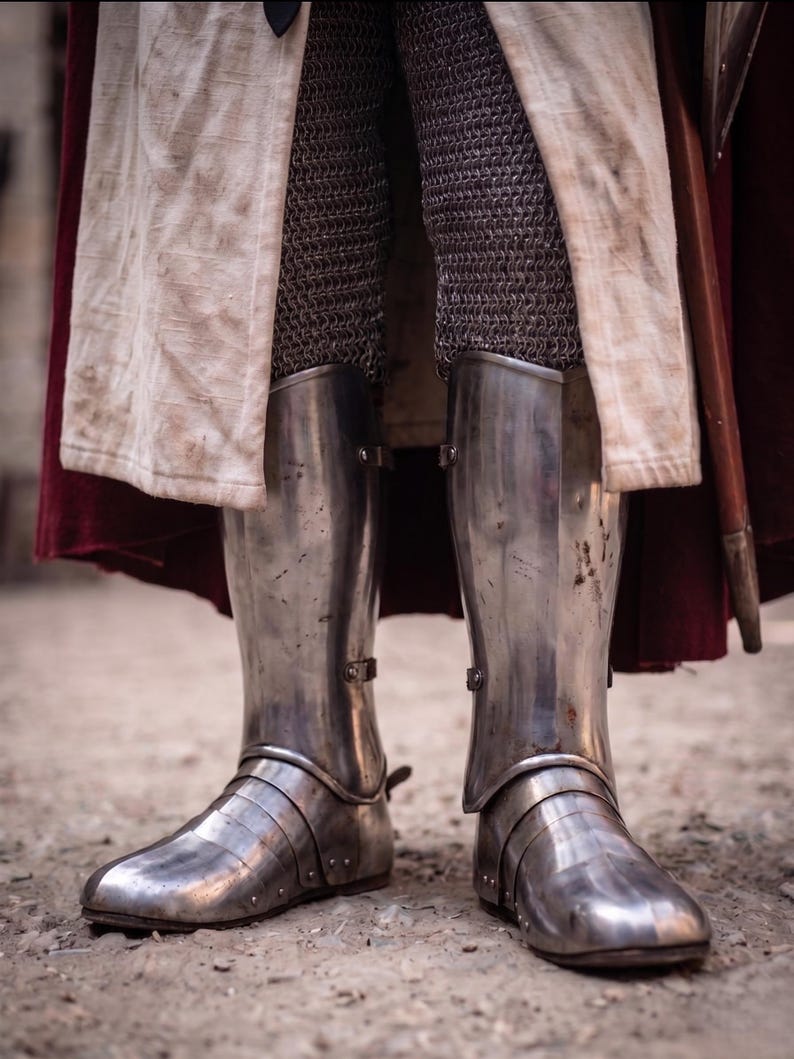 May include: Close-up of medieval steel leg armor, including greaves and sabatons. The armor is highly polished, reflecting the light. The legs are covered in chainmail. A white and red cloak is visible in the background.