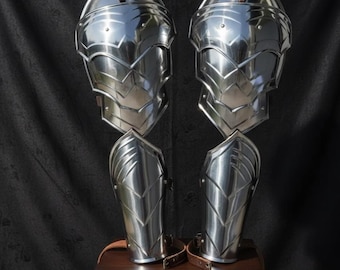 May include: A pair of polished, silver-colored metal leg armor, reflecting the light. The armor is segmented, with overlapping plates and leather straps. The pieces are displayed on a small wooden table against a dark backdrop.