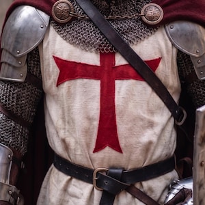 May include: A medieval knight's attire, featuring a cream-colored tunic with a large red cross, chainmail, and metal shoulder armor. A burgundy cape is draped over the shoulders, secured with decorative clasps. A black leather belt cinches the waist.