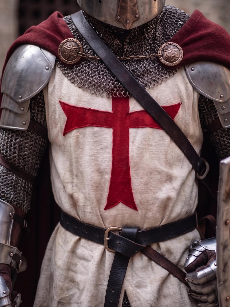 May include: A medieval knight's attire, featuring a cream-colored tunic with a large red cross, chainmail, and metal shoulder armor. A burgundy cape is draped over the shoulders, secured with decorative clasps. A black leather belt cinches the waist.