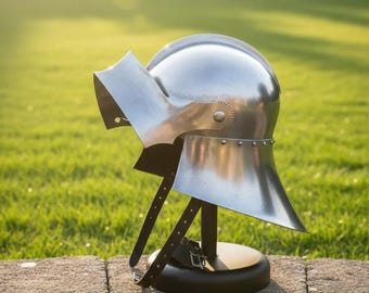 Peut inclure: Casque argenté poli avec visière et protège-nuque, présenté sur un support noir. Le casque a un sommet arrondi et un bord inférieur évasé. La visière est inclinée et de forme rectangulaire. Le support a des sangles en cuir.