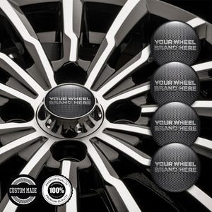 May include: Close-up of a black and silver car wheel with a central hubcap and three additional hubcaps. The hubcaps have a textured surface and the text "YOUR WHEEL BRAND HERE". The wheel has a custom made and premium quality badge.