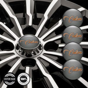 May include: Close-up of a car wheel with a black and silver spoke design. The center cap features the word "Custom" in orange script over a gray, textured background. Additional caps with the same design are visible.