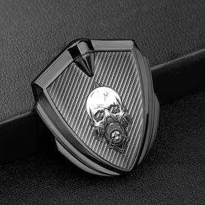 May include: A black shield-shaped emblem with a carbon fiber pattern and a skull wearing a gas mask. The emblem has a pointed top and a shiny, metallic finish.