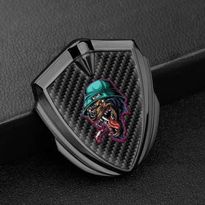 May include: Black and silver car emblem with a carbon fibre texture. The emblem features a cartoon dog wearing a green bucket hat with the word "King" on it. The dog is smoking a cigarette and has its tongue sticking out.