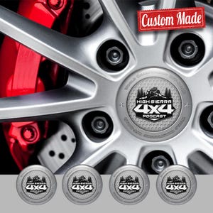 May include: Silver car wheel with black lug nuts and a red brake caliper. The wheel center features a custom-made emblem with the text "High Sierra 4x4 Podcast" and a mountain graphic. Four additional emblems are displayed below.