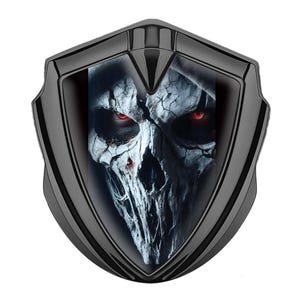 May include: A black shield-shaped emblem featuring a detailed image of a skull with glowing red eyes. The skull is depicted in shades of white and gray, with a dark background. The emblem has a glossy finish.