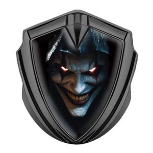 Può includere: Un emblema nero a forma di scudo con un volto di clown minaccioso con occhi rossi incandescenti e un ampio sorriso. Il volto del clown è dettagliato con toni bianchi, blu e arancioni, su uno sfondo scuro.